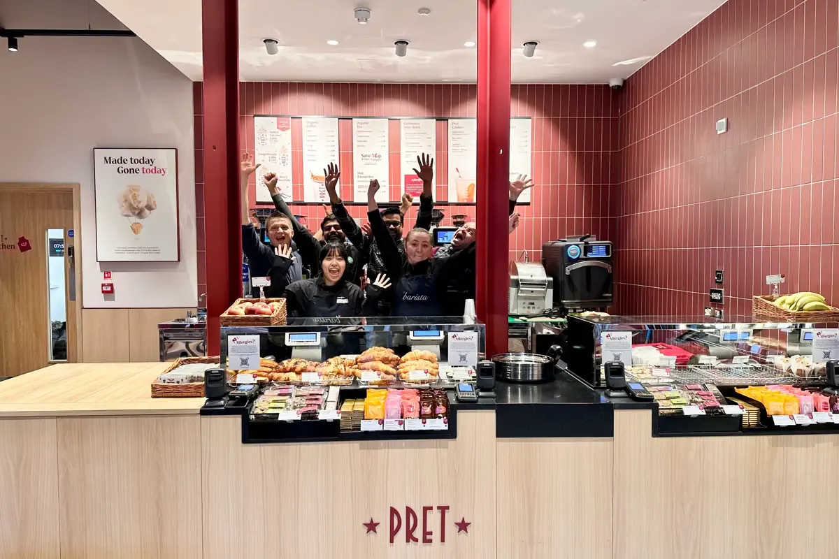 The team of a Pret A Manger shop with their arms in the air behind the pastry counter