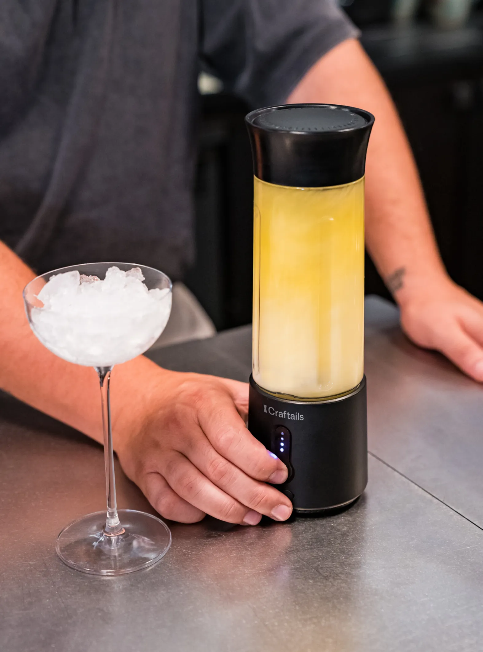 Une main pose un shaker Craftails rempli d'un cocktail jaune sur un comptoir, à côté d'une coupe remplie de glace pilée